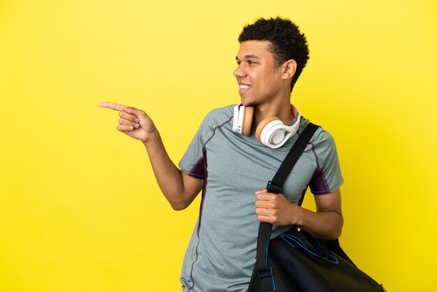 Joven deporte afroamericano con bolsa de deporte aislado sobre fondo amarillo que señala el dedo hacia el lado y presenta un producto