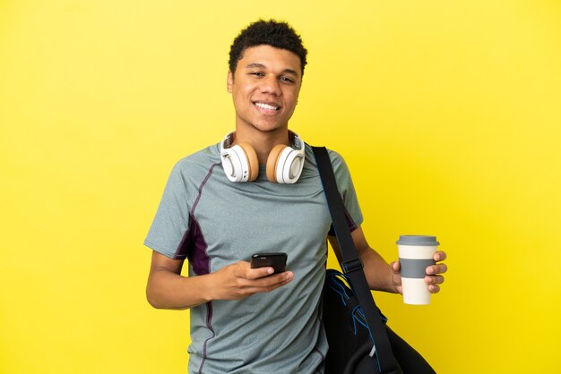 Foto joven deporte afroamericano con bolsa de deporte aislado sobre fondo amarillo sosteniendo café para llevar y un móvil