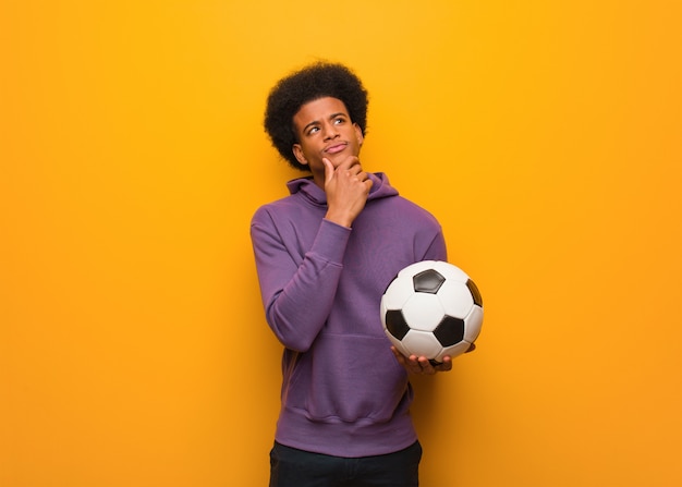 Joven deporte afroamericano hombre sosteniendo un balón de fútbol dudando y confundido