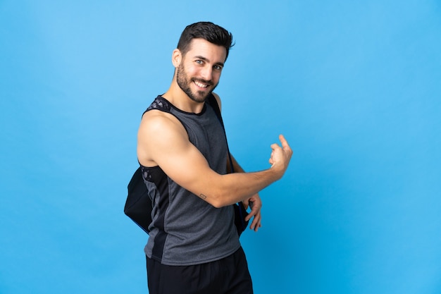 Joven deporte con bolsa de deporte aislado en la pared azul apuntando hacia atrás