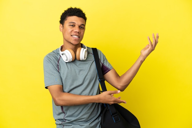 Joven deporte hombre afroamericano con bolsa de deporte aislado sobre fondo amarillo extendiendo las manos hacia el lado para invitar a venir