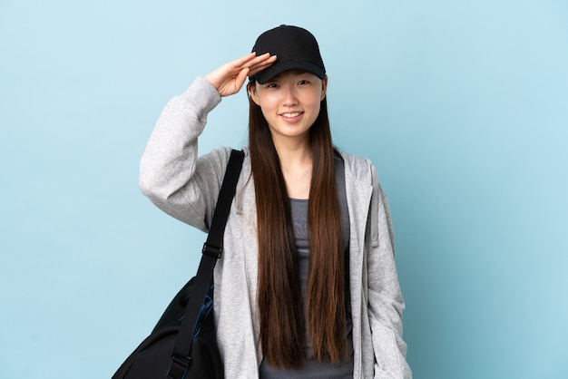 Joven deporte mujer China con bolsa de deporte sobre pared azul aislada saludando con la mano con expresión feliz