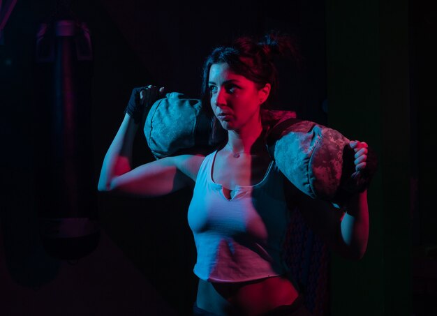 Joven deporte mujer sosteniendo una pesada bolsa de fitness para entrenamiento en luz roja azul degradado de neón sobre una pared oscura Concepto de entrenamiento funcional