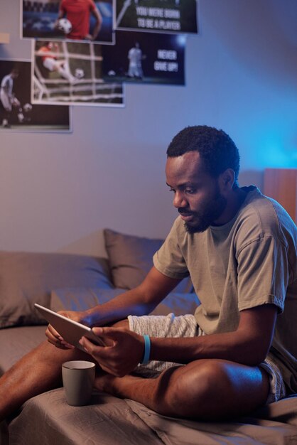 Joven deportista africano serio en ropa deportiva sentado en la cama y viendo el juego en línea en la pantalla de la tableta digital