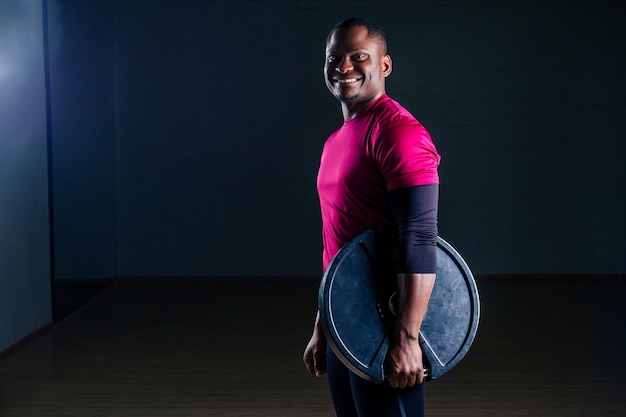 Joven deportista afroamericano en cuclillas deportista ejercicio con mancuernas camiseta uniformes deportivos ropa deportiva gimnasio de estudio de fondo negro