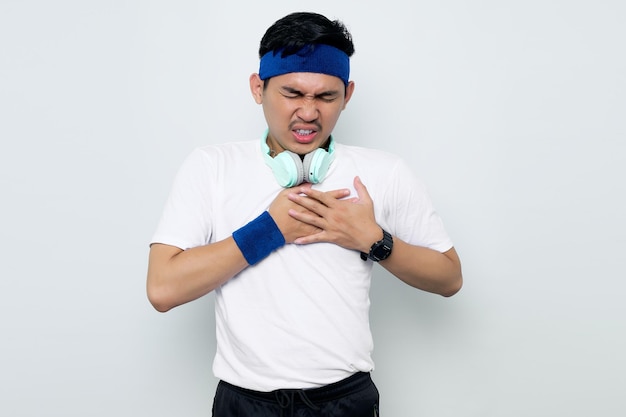 Joven deportista asiático con diadema azul y camiseta blanca con auriculares presiona una mano contra el pecho tiene un ataque al corazón que sufre de un dolor insoportable cierra los ojos aislado en el fondo blanco