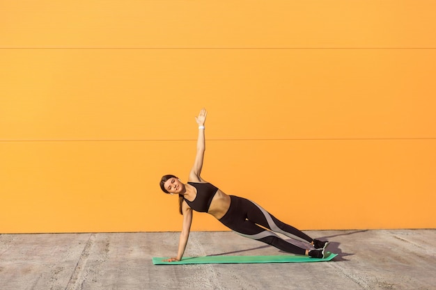 Joven deportista atractiva practicando yoga, haciendo ejercicio vasisthasana, pose de tablón lateral, haciendo ejercicio en alfombra verde, usando ropa deportiva negra, al aire libre, fondo naranja, deporte y concepto saludable