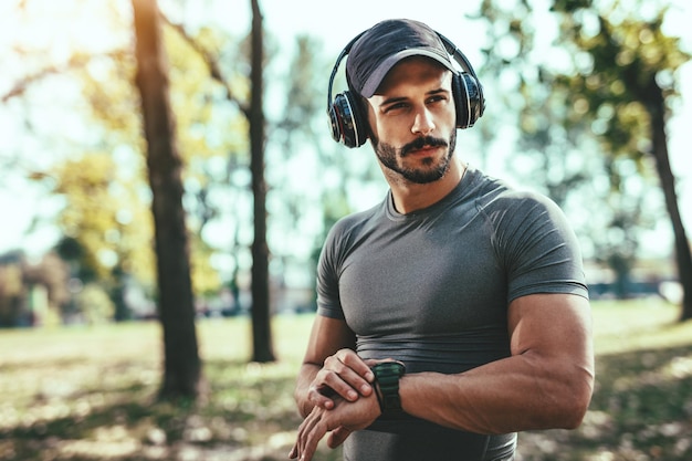 Joven deportista con auriculares descansa después de entrenar en el parque y mira el cronómetro.