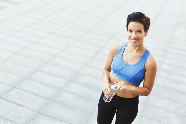 Una joven deportista con auriculares está tomando un descanso, bebiendo agua después de hacer ejercicio en la ciudad, mirando hacia arriba y sonriendo a la cámara, copiando espacio