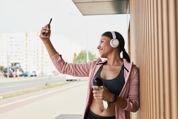Foto joven deportista en auriculares inalámbricos sonriendo a la cámara mientras hace retrato selfie en teléfono móvil ...