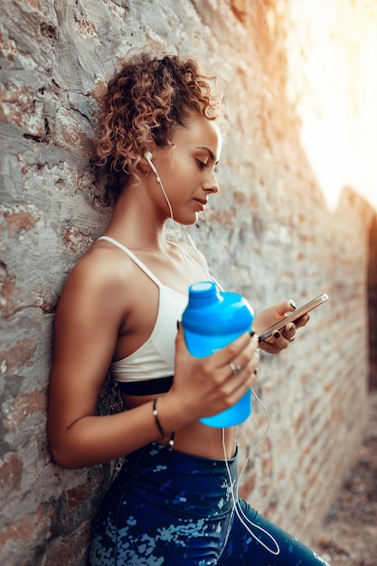 Joven deportista con auriculares tomando un descanso después de un duro entrenamiento y usando el teléfono móvil.