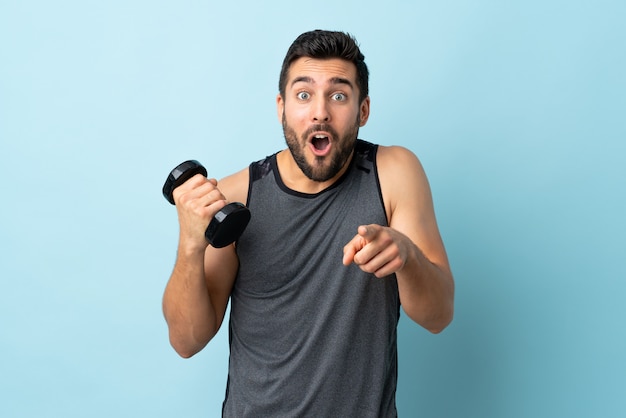 Joven deportista con barba haciendo levantamiento de pesas sorprendido y apuntando al frente