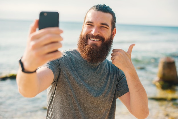 Joven deportista barbudo tomando selfie con smartphone cerca del océano y mostrando el pulgar hacia arriba