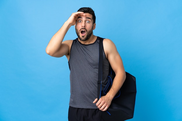 Joven deportista con bolsa de deporte aislado en la pared azul haciendo gesto de sorpresa mientras mira hacia el lado