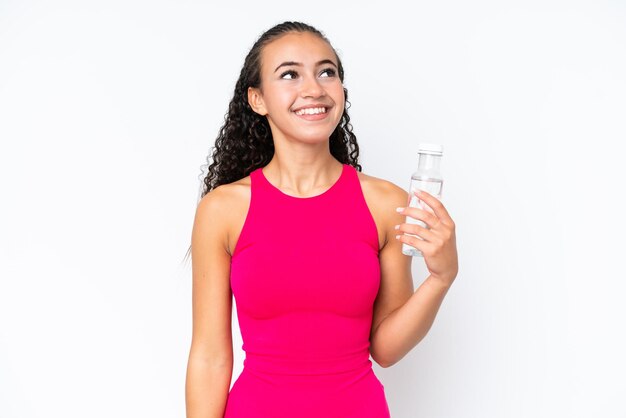 Joven deportista con una botella de agua aislada en blanco mirando hacia arriba mientras sonríe