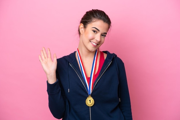 Joven deportista brasileña con medallas aisladas de fondo rosa saludando con la mano con expresión feliz