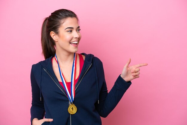 Joven deportista brasileña con medallas aisladas de fondo rosa señalando con el dedo al costado y presentando un producto