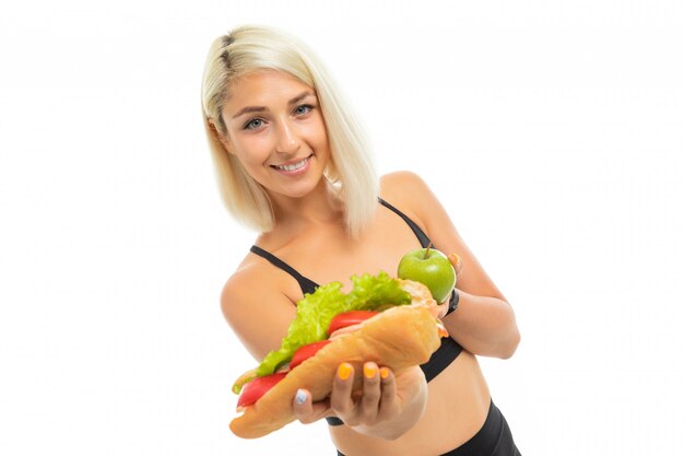 Una joven deportista con cabello rubio sostiene una manzana verde y un sándwich