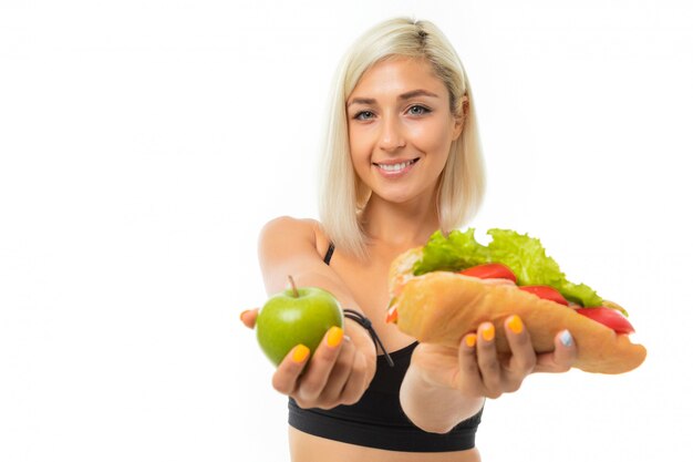 Una joven deportista con cabello rubio en un top deportivo negro y calzas negras sostiene una manzana verde y un sándwich.