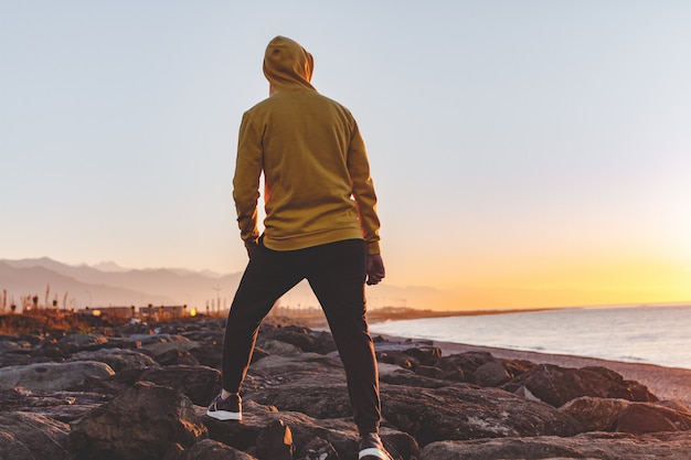 Joven deportista en el capó haciendo un salto sobre las rocas en el fondo del mar y las montañas al atardecer