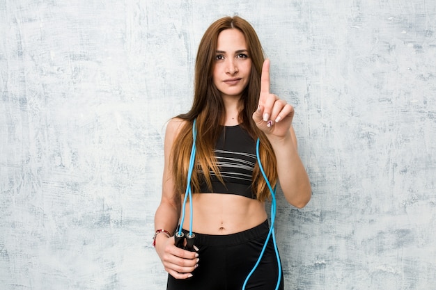 Joven deportista caucásica sosteniendo una cuerda para saltar mostrando el número uno con el dedo.