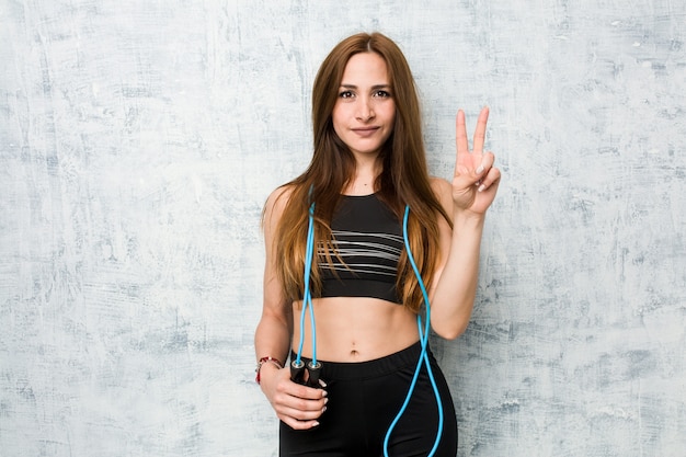 Joven deportista caucásica sosteniendo una cuerda para saltar mostrando el número dos con los dedos