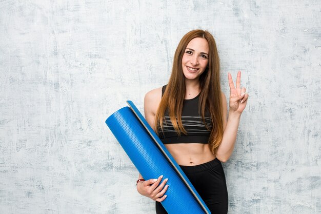 Joven deportista caucásica sosteniendo una estera que muestra el número dos con los dedos.