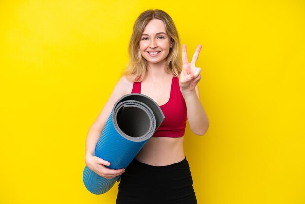 Joven deportista caucásica yendo a clases de yoga mientras sostiene una alfombra aislada en un fondo amarillo sonriendo y mostrando el signo de la victoria