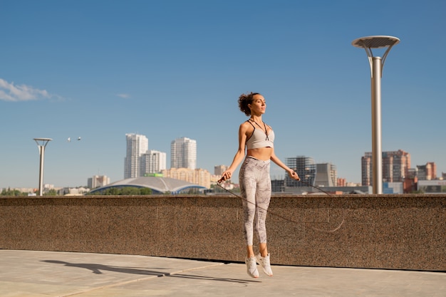 Joven deportista delgada en ropa deportiva haciendo ejercicio con saltar la cuerda mientras entrena en un entorno urbano contra la orilla del río y el cielo azul