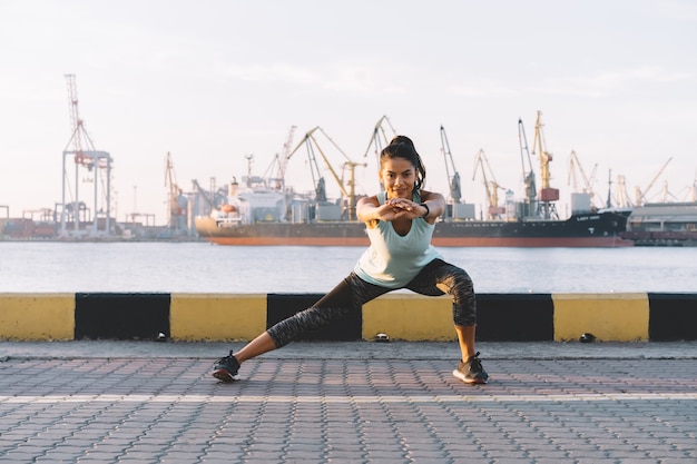 Joven deportista estirando y preparándose para correr cerca del puerto marítimo.