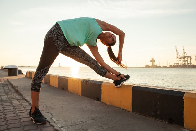 Joven deportista estirando y preparándose para correr cerca del puerto marítimo.