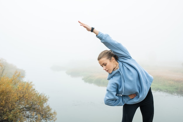 Foto joven deportista en forma con sudadera con capucha azul y leggins negros estirando su brazo izquierdo sobre sí misma mientras hace curvas laterales en un entorno natural