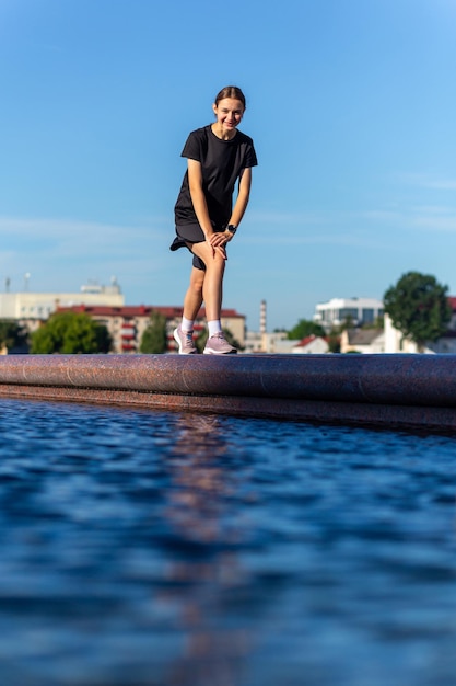 Joven deportista y en forma vestida de negro estirándose después del entrenamiento cerca de la fuente pública en el parque urbano Fitness deporte jogging urbano y concepto de estilo de vida saludable