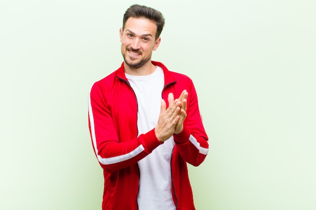 Joven deportista guapo o monitor sintiéndose feliz y exitoso, sonriendo y aplaudiendo, saludando con un aplauso contra la pared verde