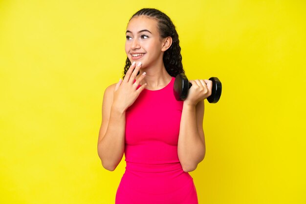 Joven deportista haciendo levantamiento de pesas aislado de fondo amarillo mirando hacia arriba mientras sonríe