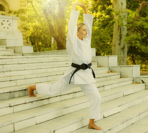 Joven deportista con un kimono blanco con cinturón negro hace un calentamiento en las escaleras antes de entrenar. Artes marciales, defensa personal