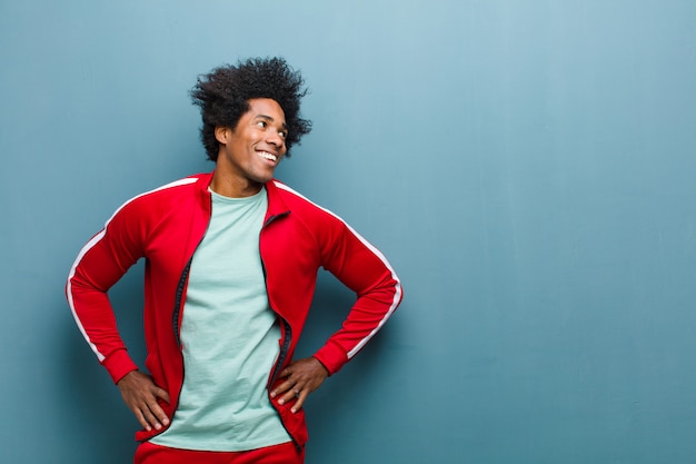 Joven deportista negro que parece feliz, alegre y confiado, sonriendo con orgullo y mirando al lado con ambas manos en las caderas