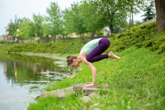 Una joven deportista practica yoga en un césped verde junto al río, el yoga ataca la postura. Meditación y unidad con la naturaleza.