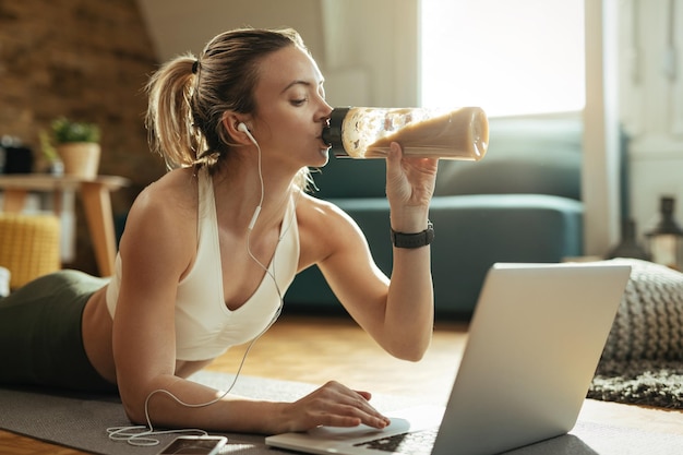 Joven deportista relajándose en el suelo y bebiendo batido mientras usa una laptop en casa