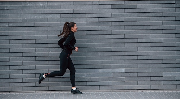 Joven deportista en ropa deportiva negra corriendo al aire libre cerca de la pared gris