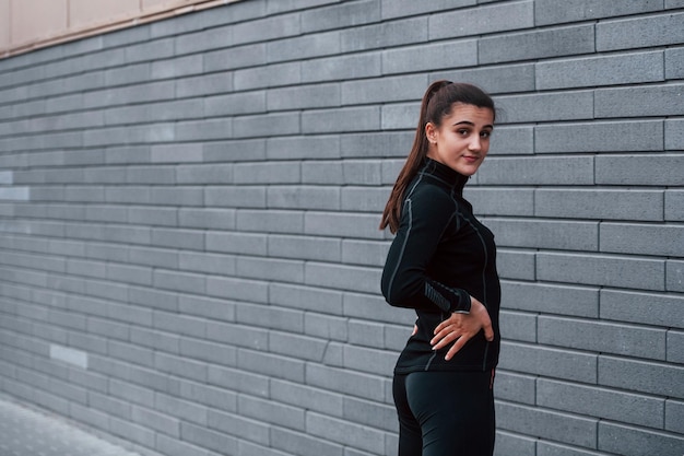 Joven deportista en ropa deportiva negra de pie al aire libre cerca de la pared gris