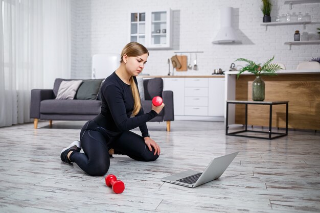 Joven deportista en ropa de fitness en una casa moderna usando lecciones en línea de un sitio de fitness en una computadora portátil y haciendo ejercicio en casa. Deportes en casa durante la cuarentena.