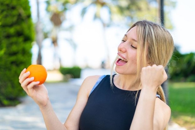 Joven deportista rubia sosteniendo una naranja al aire libre celebrando una victoria