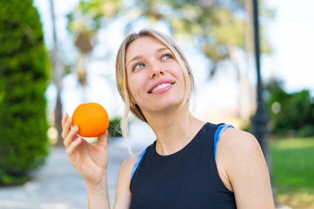 Joven deportista rubia sosteniendo una naranja al aire libre mirando hacia arriba mientras sonríe