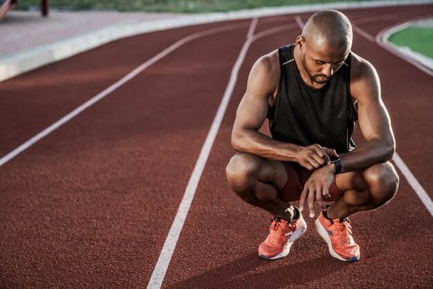 Joven deportista sentado en la pista de atletismo y mirando el reloj inteligente