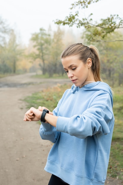 Foto joven deportista seria en sudadera con capucha azul mirando el reloj de pulsera antes de entrenar mientras está de pie frente a la cámara contra el camino del bosque