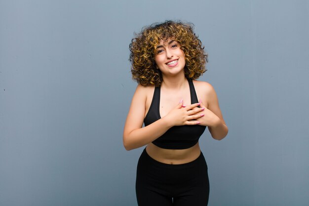 Joven deportista sintiéndose romántica, feliz y enamorada, sonriendo alegremente y cogidos de la mano cerca del corazón contra la pared gris