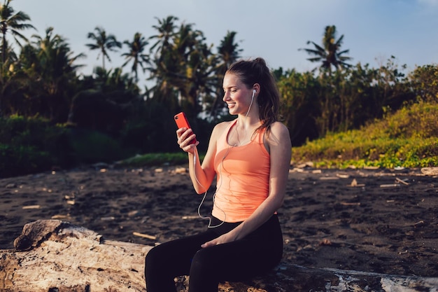 Joven deportista sonriente escuchando música y usando un teléfono inteligente contra el río y las palmeras