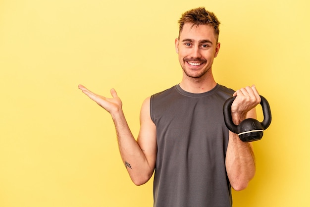 Joven deportista sosteniendo una pesa aislada en un fondo amarillo mostrando un espacio de copia en una palma y sosteniendo otra mano en la cintura.