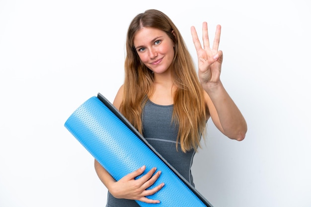 Joven deportista yendo a clases de yoga mientras sostiene una colchoneta feliz y cuenta tres con los dedos